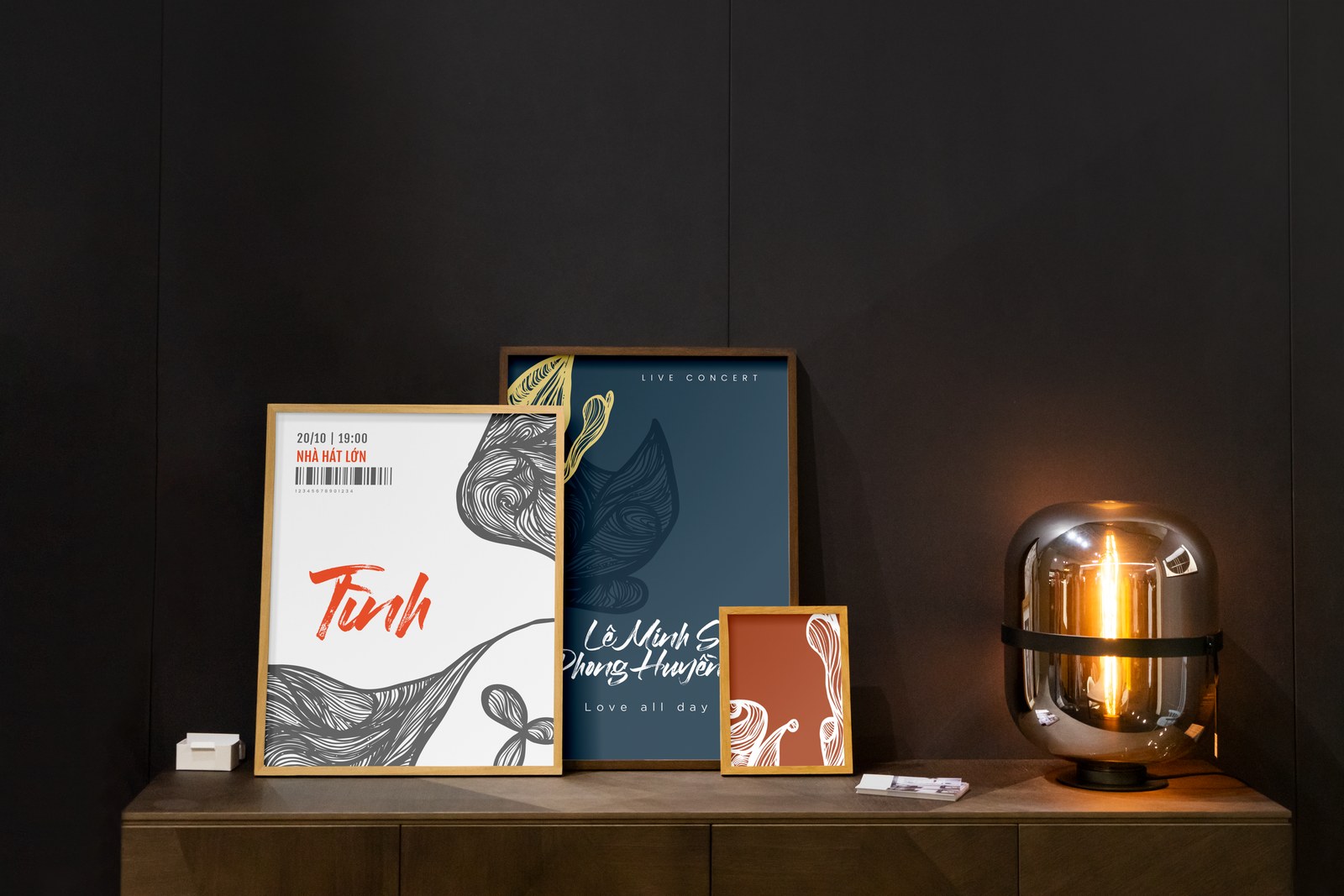 Framed posters for the album Tình arranged on a wooden cabinet beside a table lamp