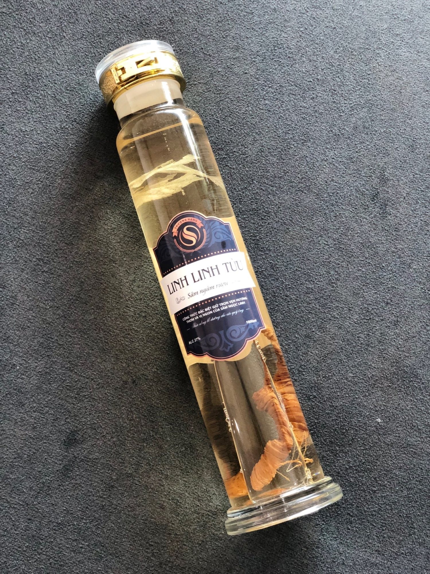 A Linh Linh Tửu glass bottle with a gold stopper, navy label on the body, the ginseng root clearly visible inside