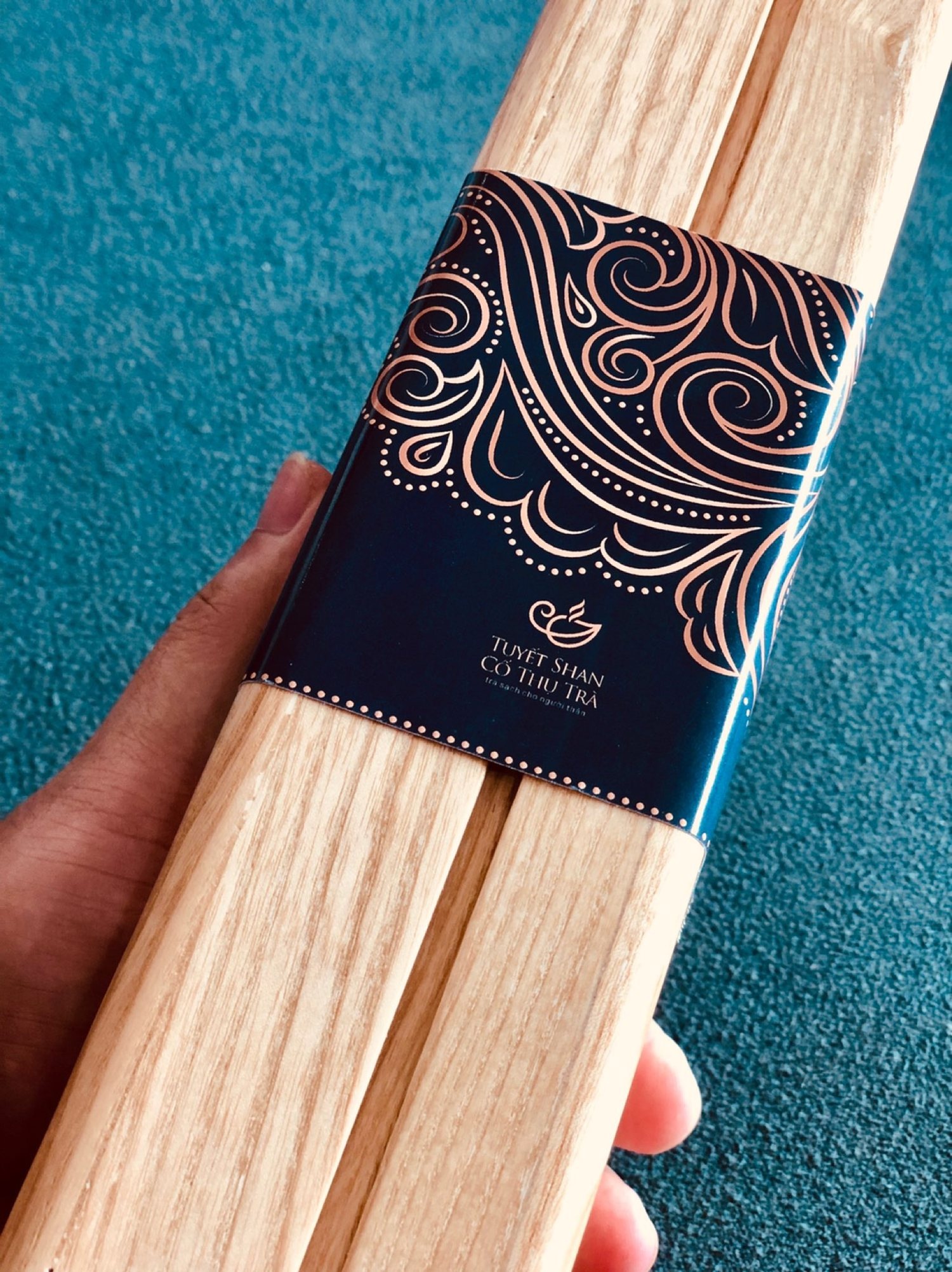A hand holding a LinhLinhSam pine box wrapped in a navy paper band printed with a gold cloud pattern