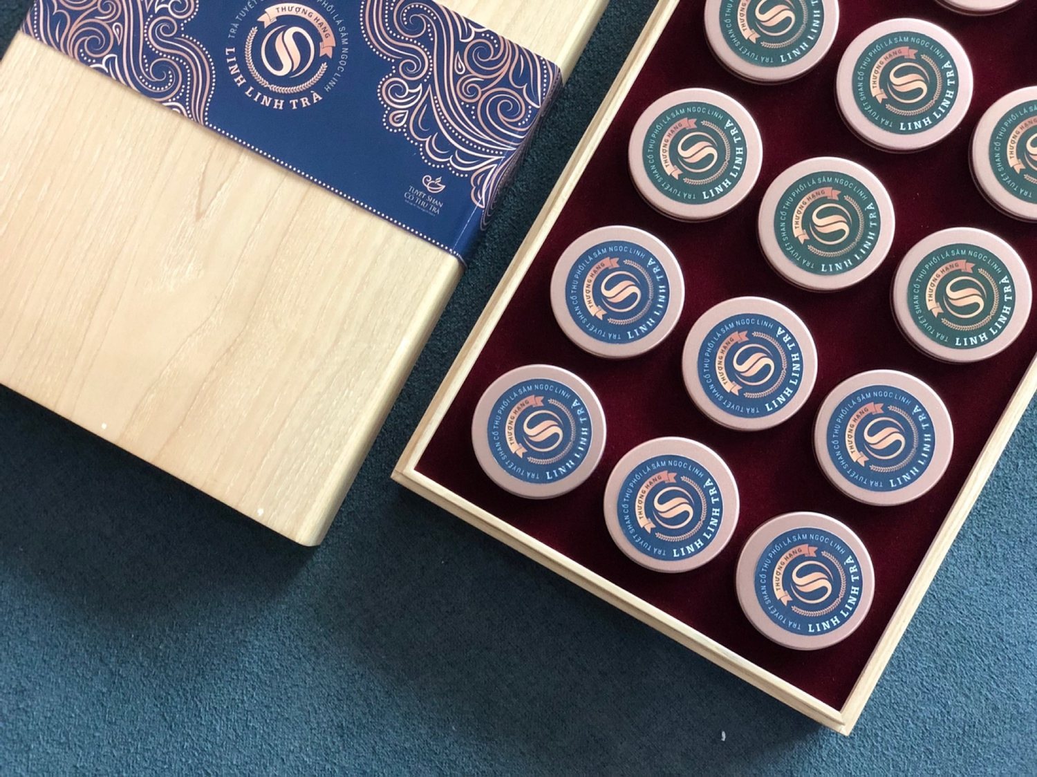 An open pine box with twelve LinhLinhSam ginseng jars seated in deep plum velvet, the navy paper band with its gold cloud pattern resting alongside