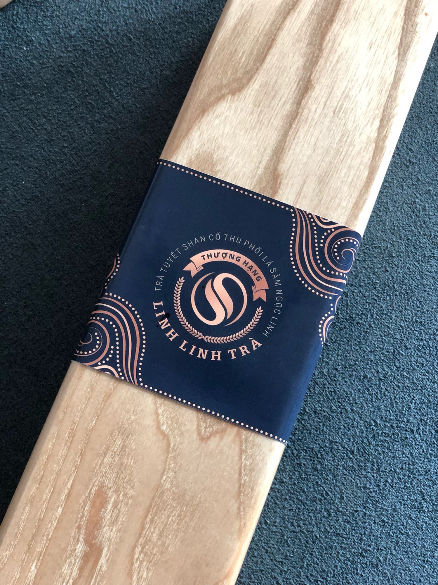 A navy paper band wrapped diagonally around the Linh Linh Trà pine box, the round S seal centred in its band of antique gold type