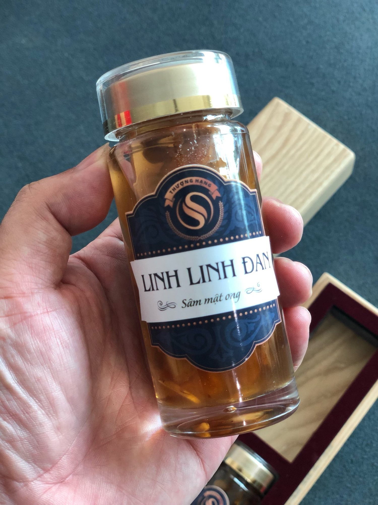 A hand holding a Linh Linh Đan glass jar, its navy label carrying the round premium mark above a white band reading ginseng in honey